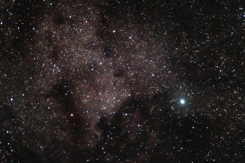 This image captures a dense expanse of the Milky Way, filled with thousands of stars in varying hues of blue, white, and orange. Dark nebulae and dust clouds weave through the starfield, creating subtle silhouettes that add depth and contrast to the cosmic scene. A particularly bright star shines prominently on the right side, drawing the eye amid the vast, intricate beauty of our galaxy.